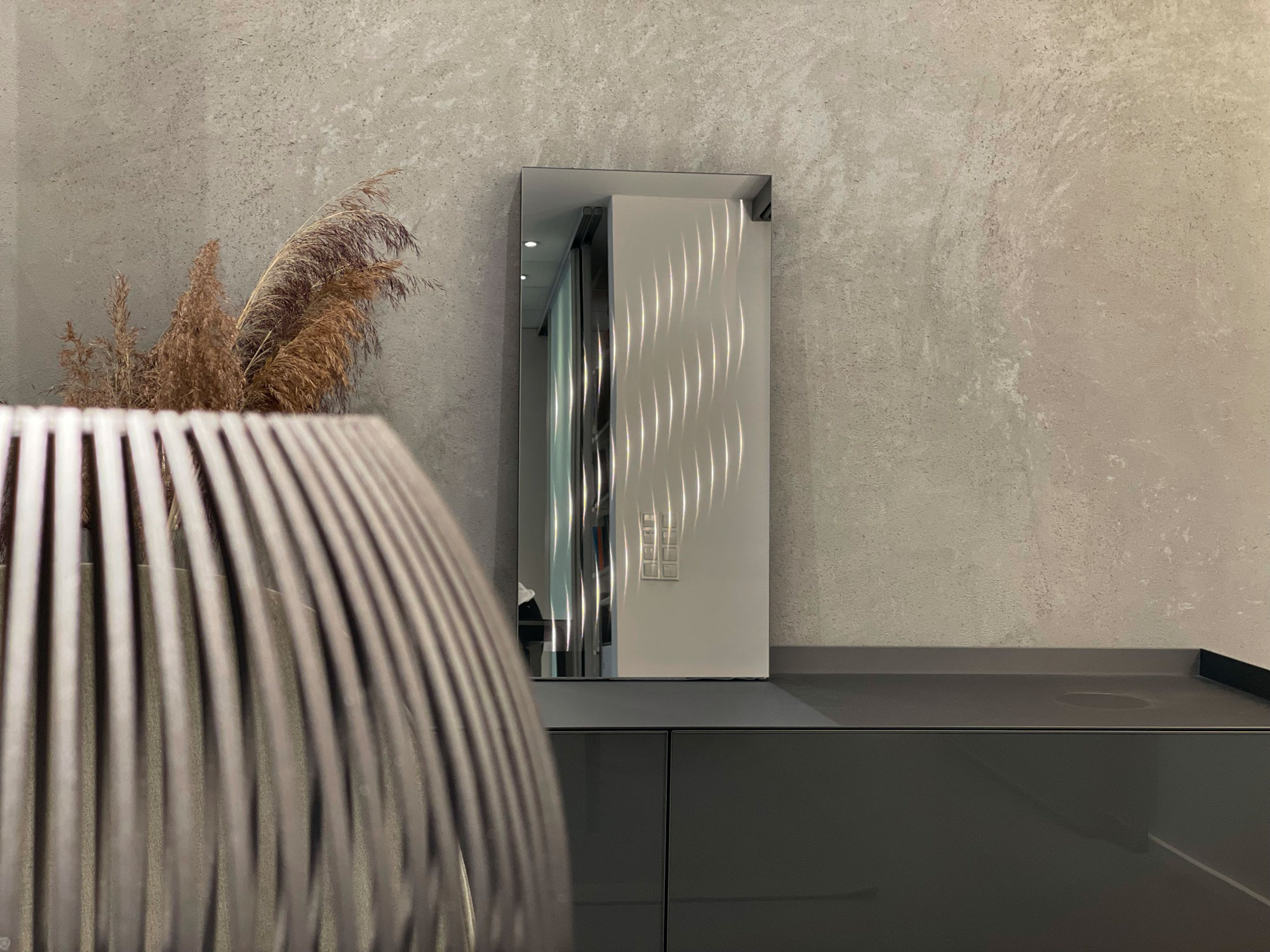 Der Lichtspiegel Ambiloom® Mirror 800 auf einem Sideboard und vor einer grauen Betonwand